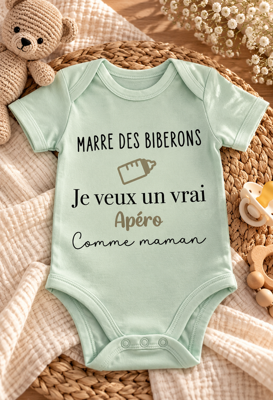 Body bébé vert menthe à manches courtes personnalisé « Marre des biberons, je veux un vrai apéro comme maman », avec motif biberon beige, présenté sur un tapis tressé avec un ourson.